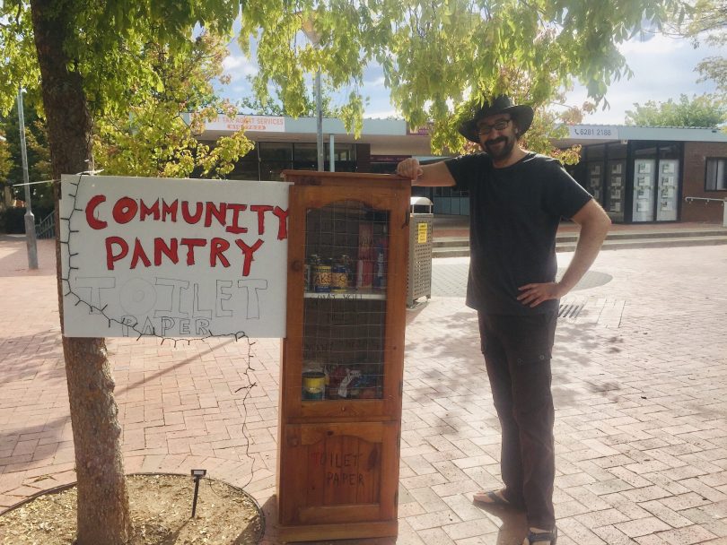 Dan Nahum standing next to a large wooden cupboard with glass front and canned food visible inside. A sign next to the cupboard reads "Community Pantry"