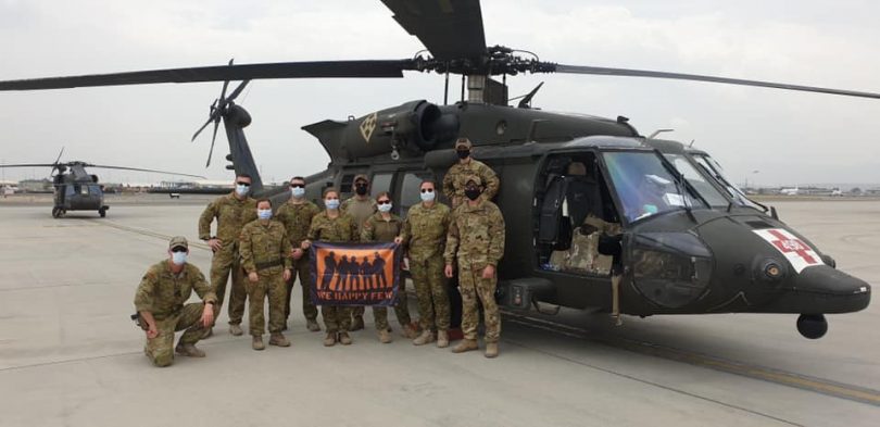 Medevac team holding We Happy Few Wines flag in Afghanistan in front of helicopter.