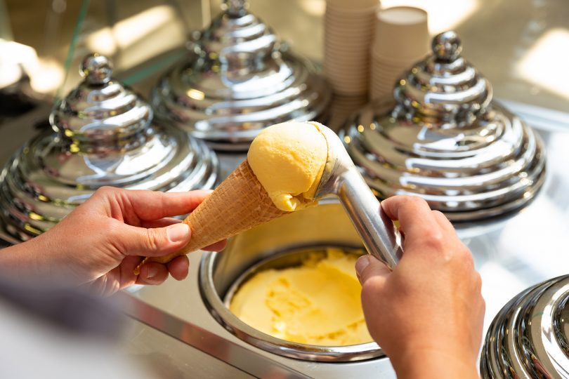 Hands use a scoop to place an orange coloured gelato onto a cone.