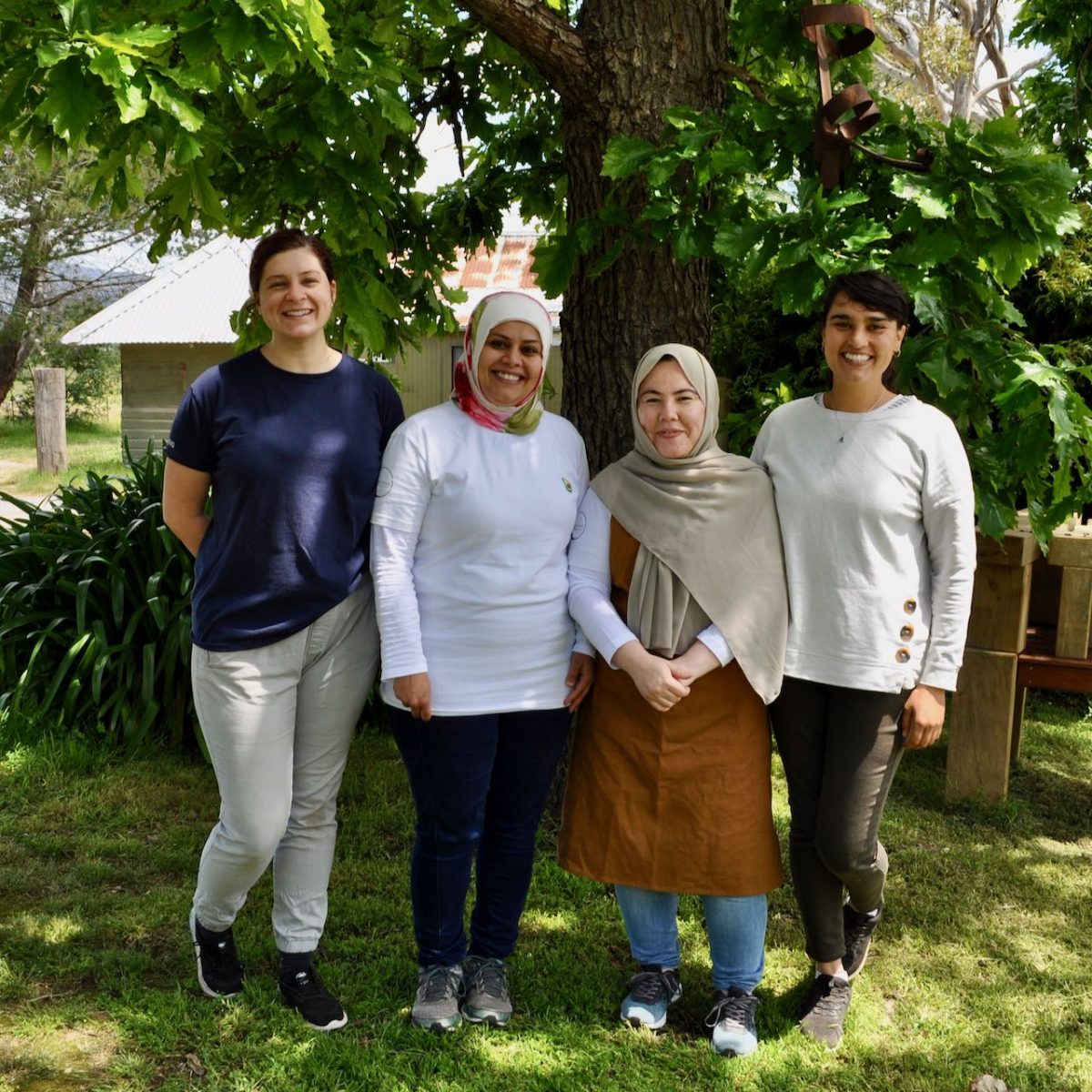 Four smiling women, two wearing headscarves, stand under a tree