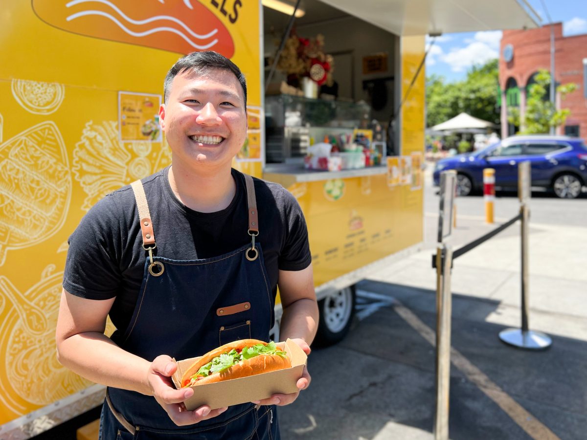 smiling man holds a banh mi in front of yellow food truck