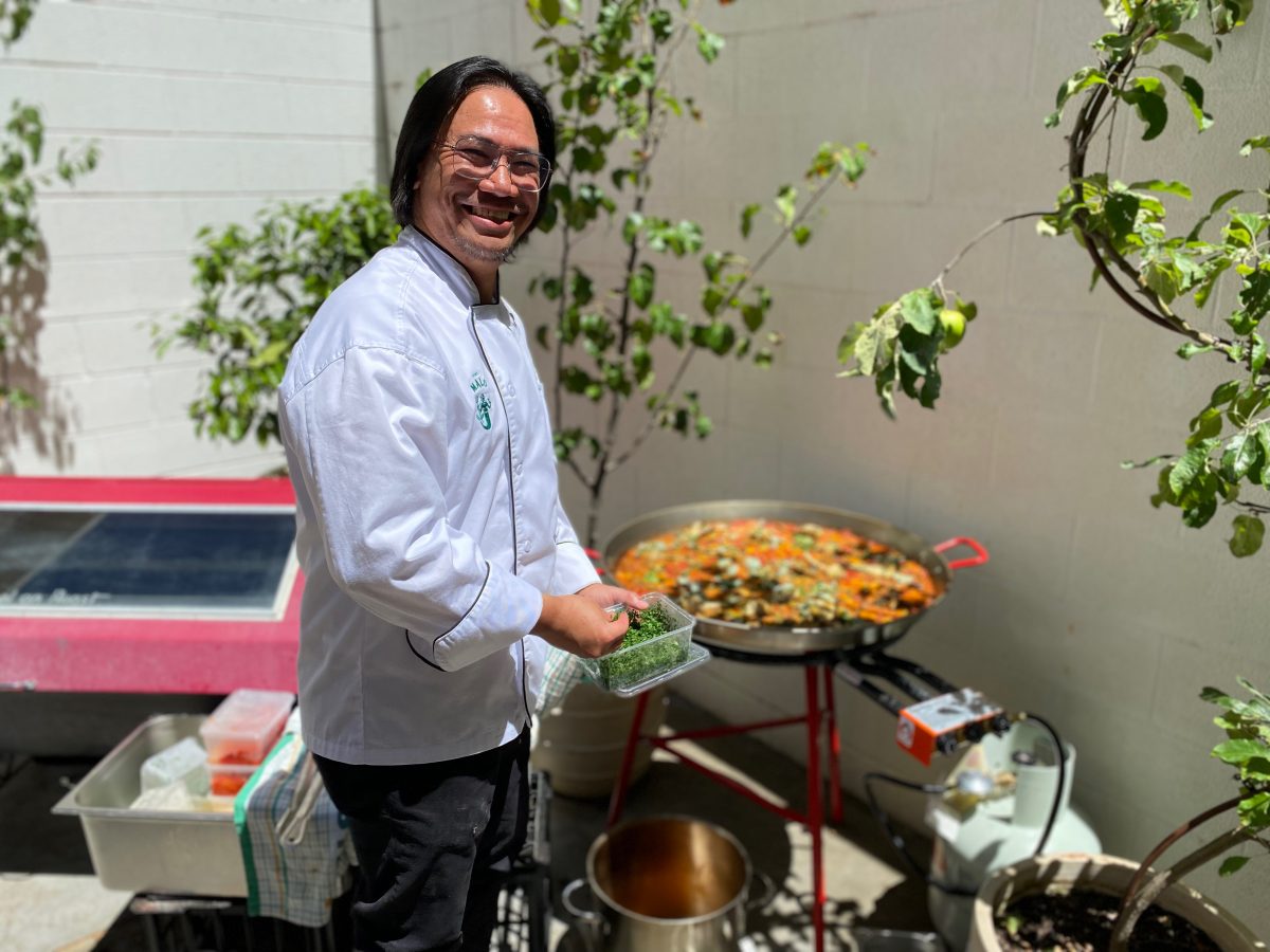 John Santos in chef's whites smiles while garnishing large pan of paella