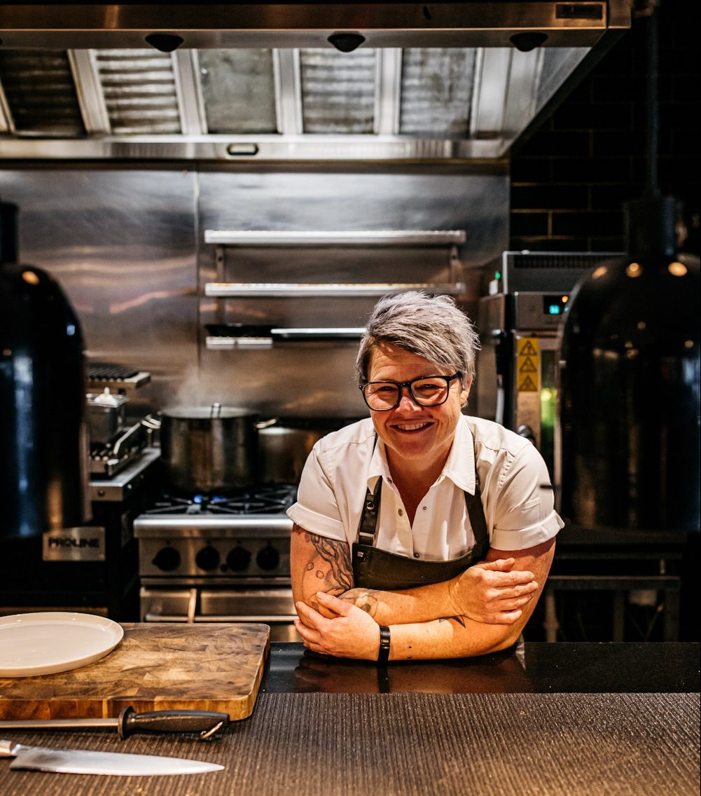 Mel, a chef in shortsleeves and apron, leans on the kitchen pass and smiles at the camera.