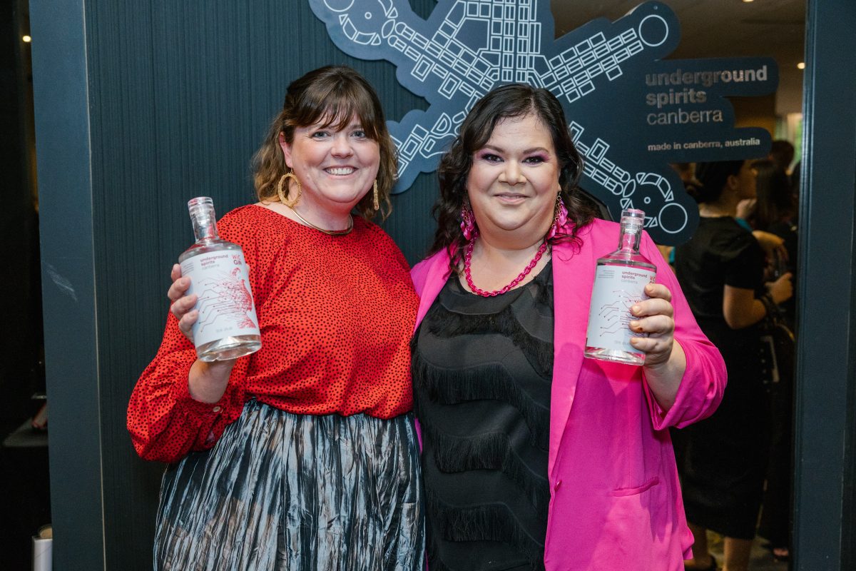 Two women, one in red and one in pink smile while holding bottles of gin