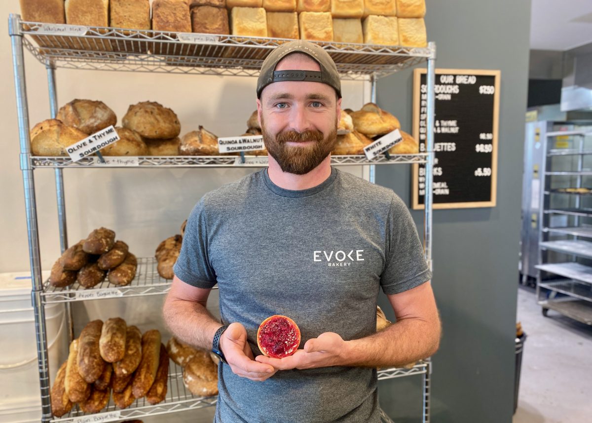 Tom holds a small red tart and stands in front of a rack of baked goods. 