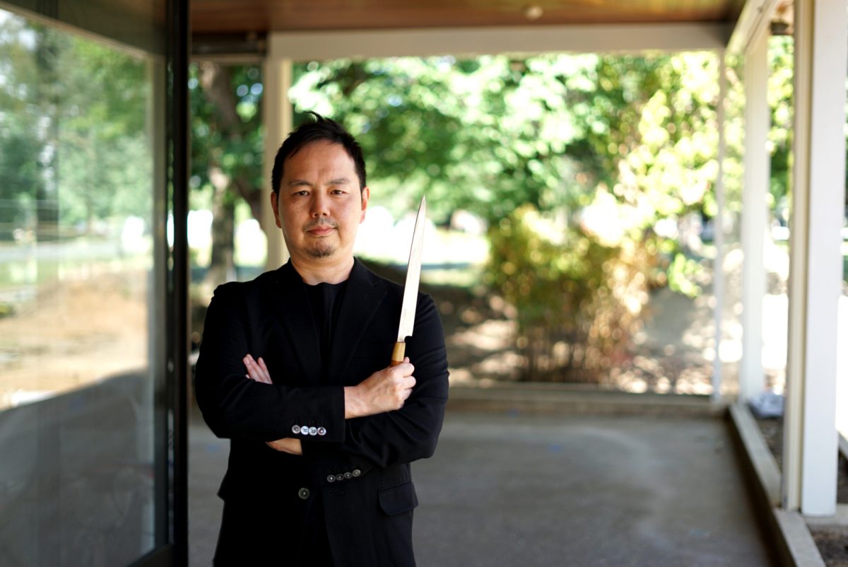 Japanese chef wearing black crosses his arms while holding chefs knife.