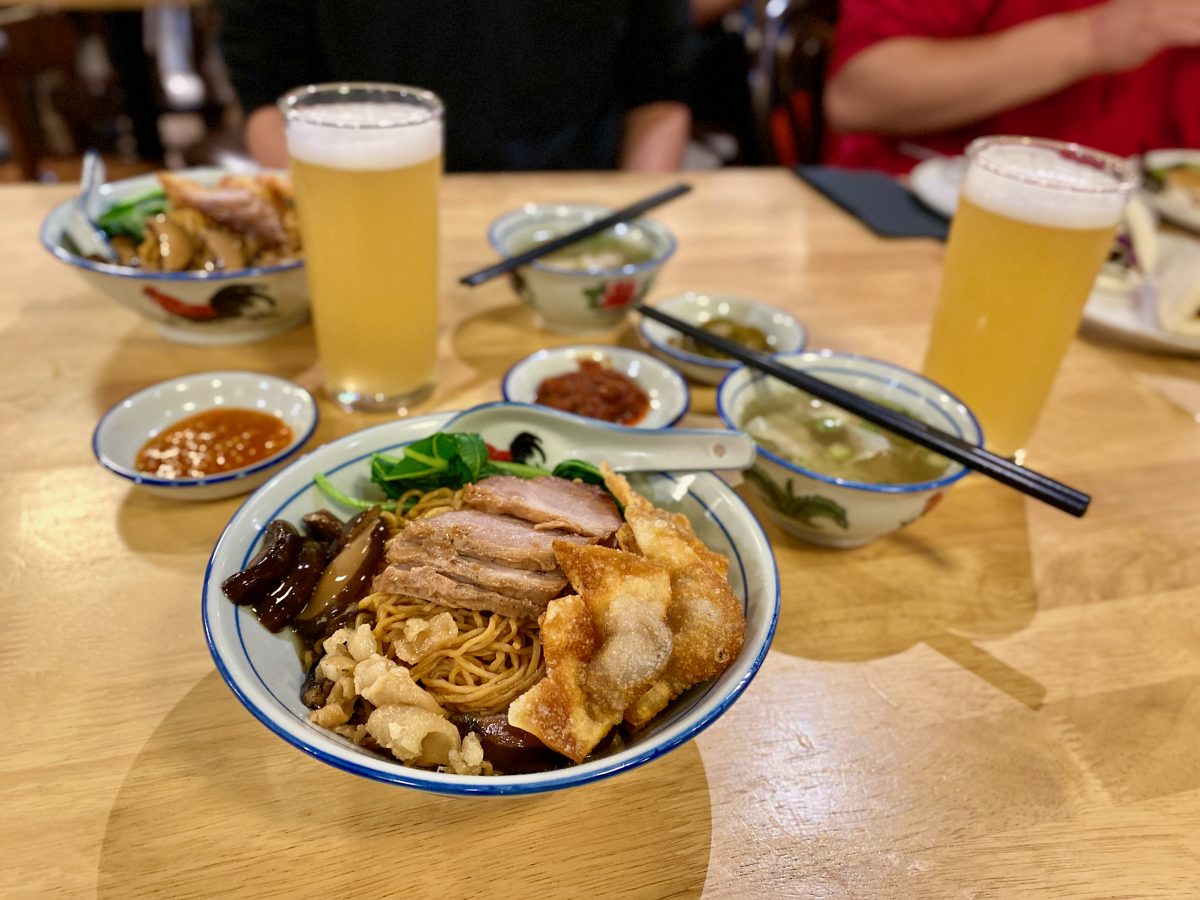 bowl of noodles with fried wonton, meat, mushrooms and greens. There are other dishes and glasses of beer in the background.