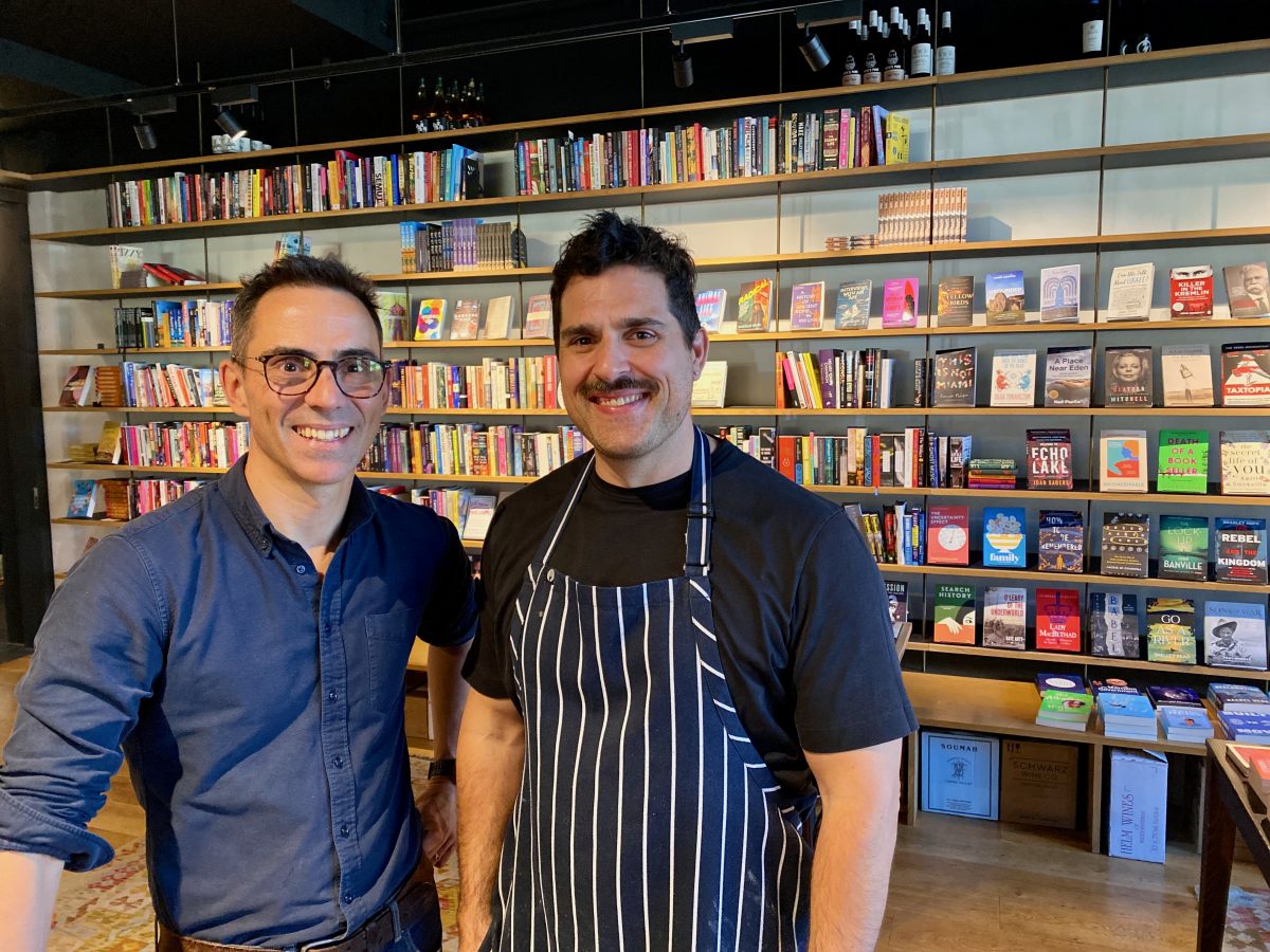 two men, one wearing an apron, smile in a bookshop