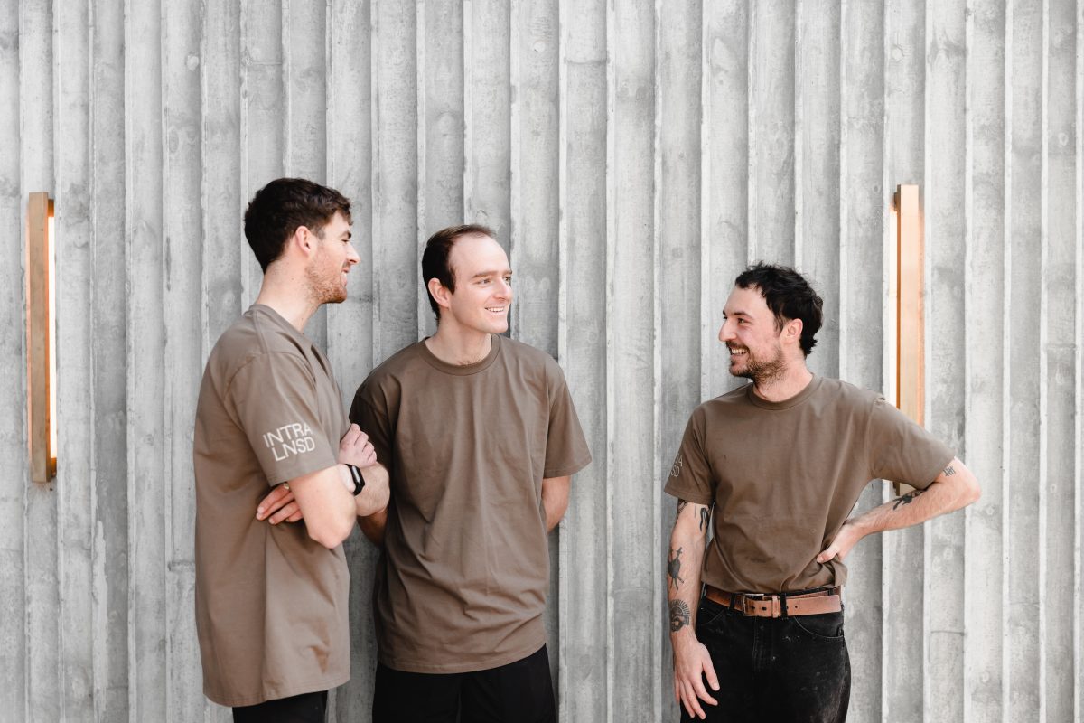 Three men in tan t-shirts pose naturally in front of textured wall