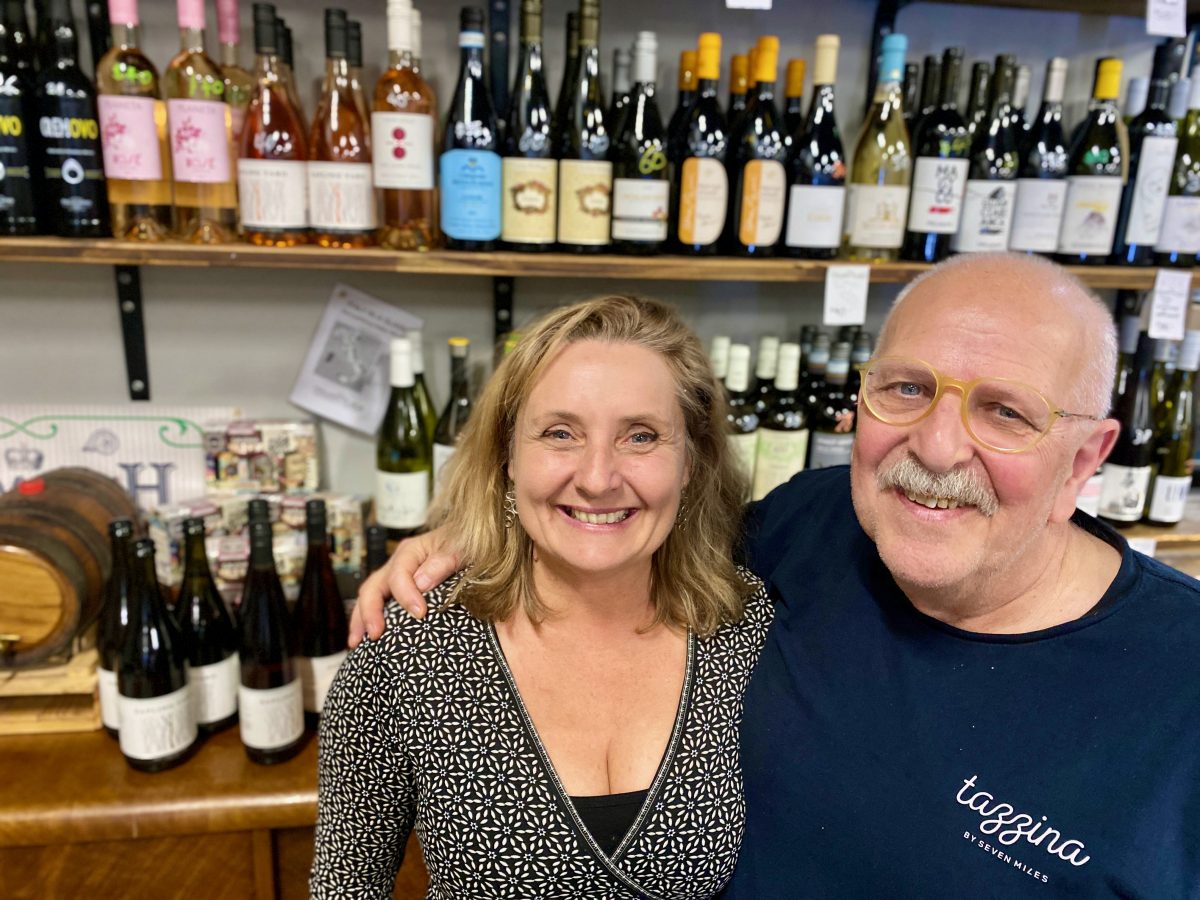 Two people smile in front of a shelf of wine