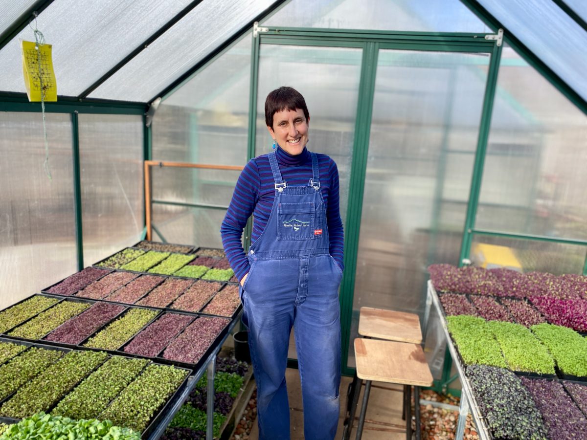 Fiona wears overalls and stands in a greenhouse surrounded by microgreens