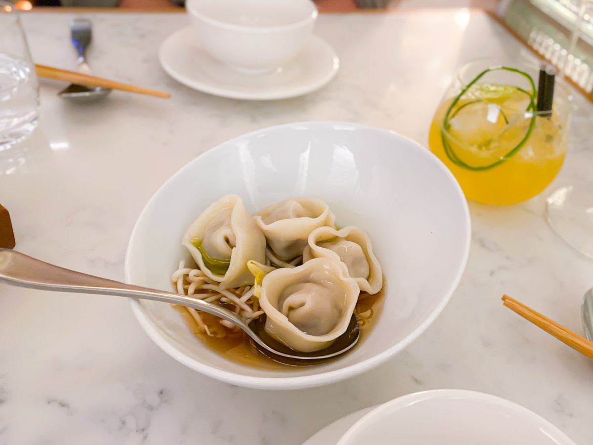 Asymmetrical bowl with four dumplings in broth, cocktail in the background