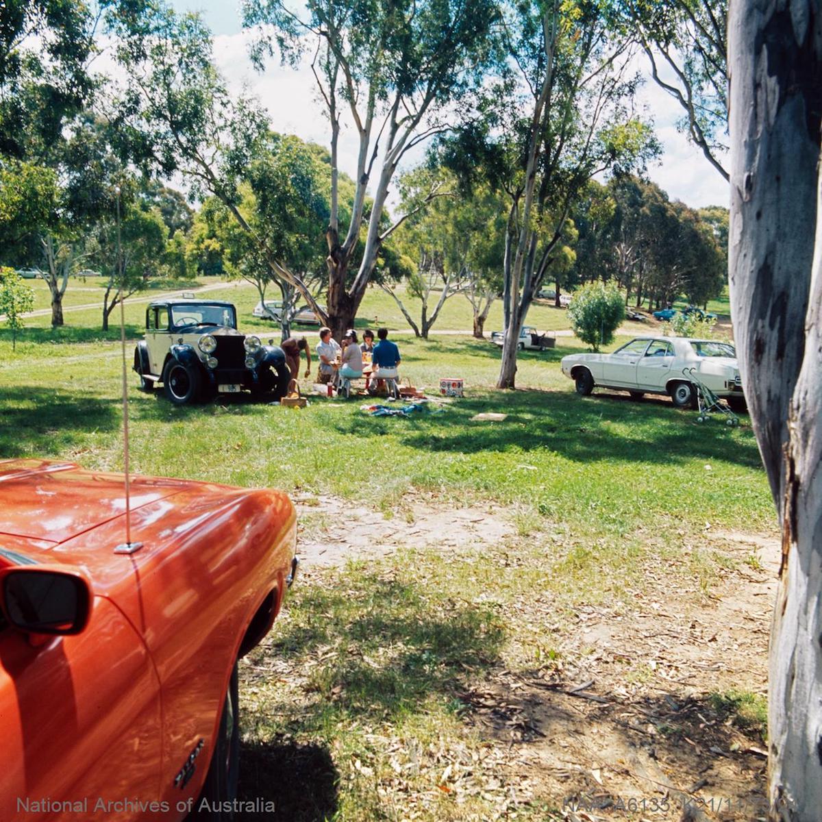 Picture shows a family picnicking at Weston Park. They are sitting at a picnic table under some shady eucalyptus trees. Nearby is a vintage car.