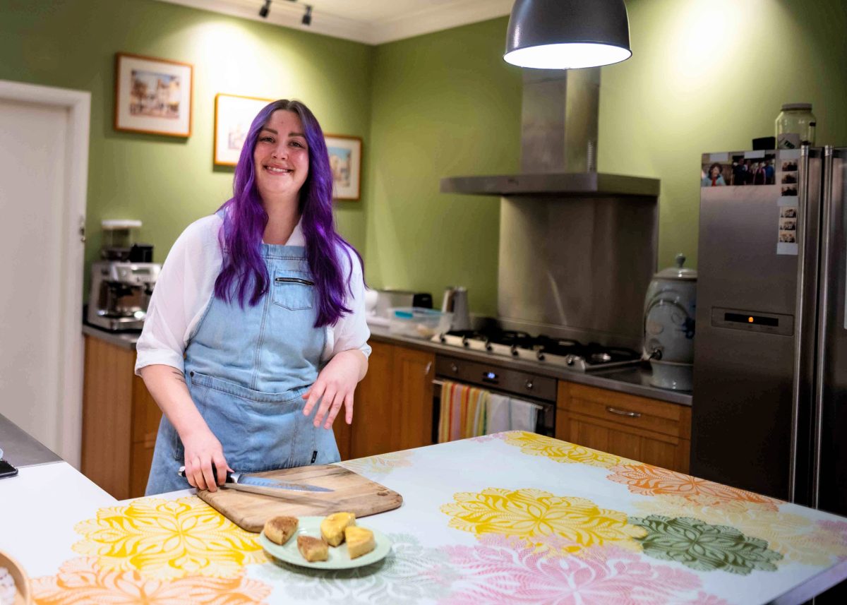 a woman with purple hair standing in a kitche with a chopping board.