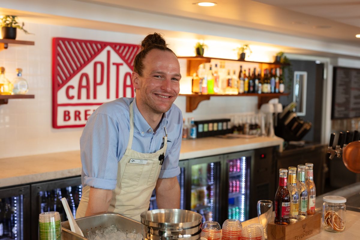 A man in apron and top-knot leaning over the bar and smiling