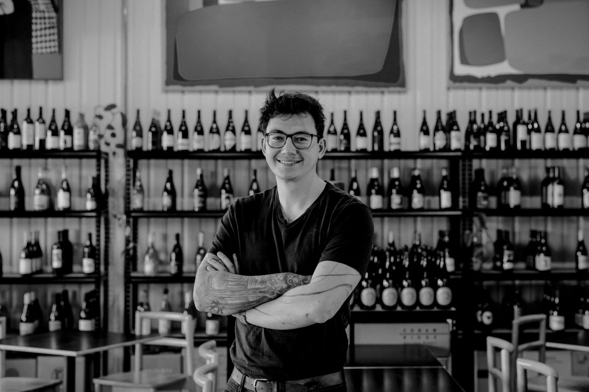 Black and white photo of Reece standing in front of a shelf of wines. 