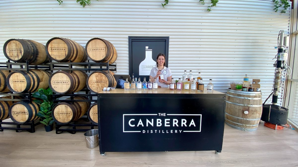 Woman stands behind table with many bottles of spirits. A sign reads "The Canberra Distillery"