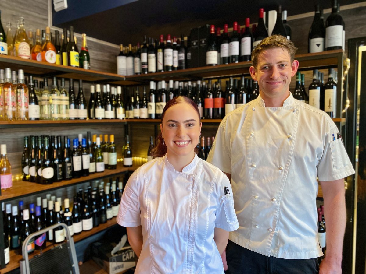 Deb and Keaton wear chefs white and smile in front of a shelf full of wine
