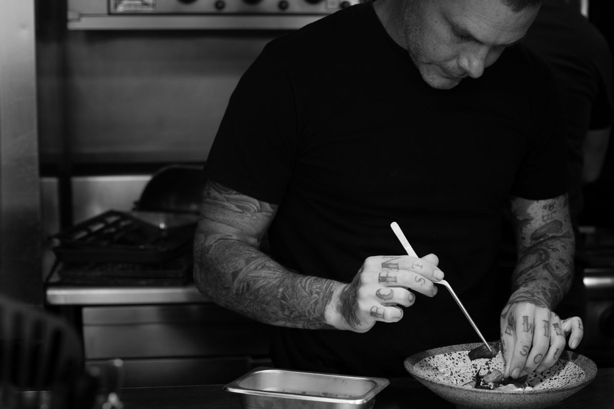 A black and white photo of a chef with tattoos plating up a dish