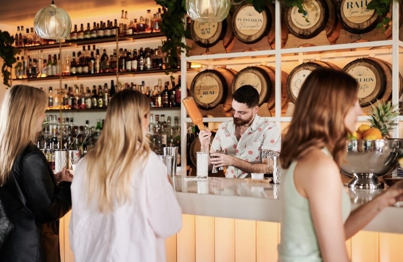 A man pouring a shot behind a bar as three women wait, their backs to the camera