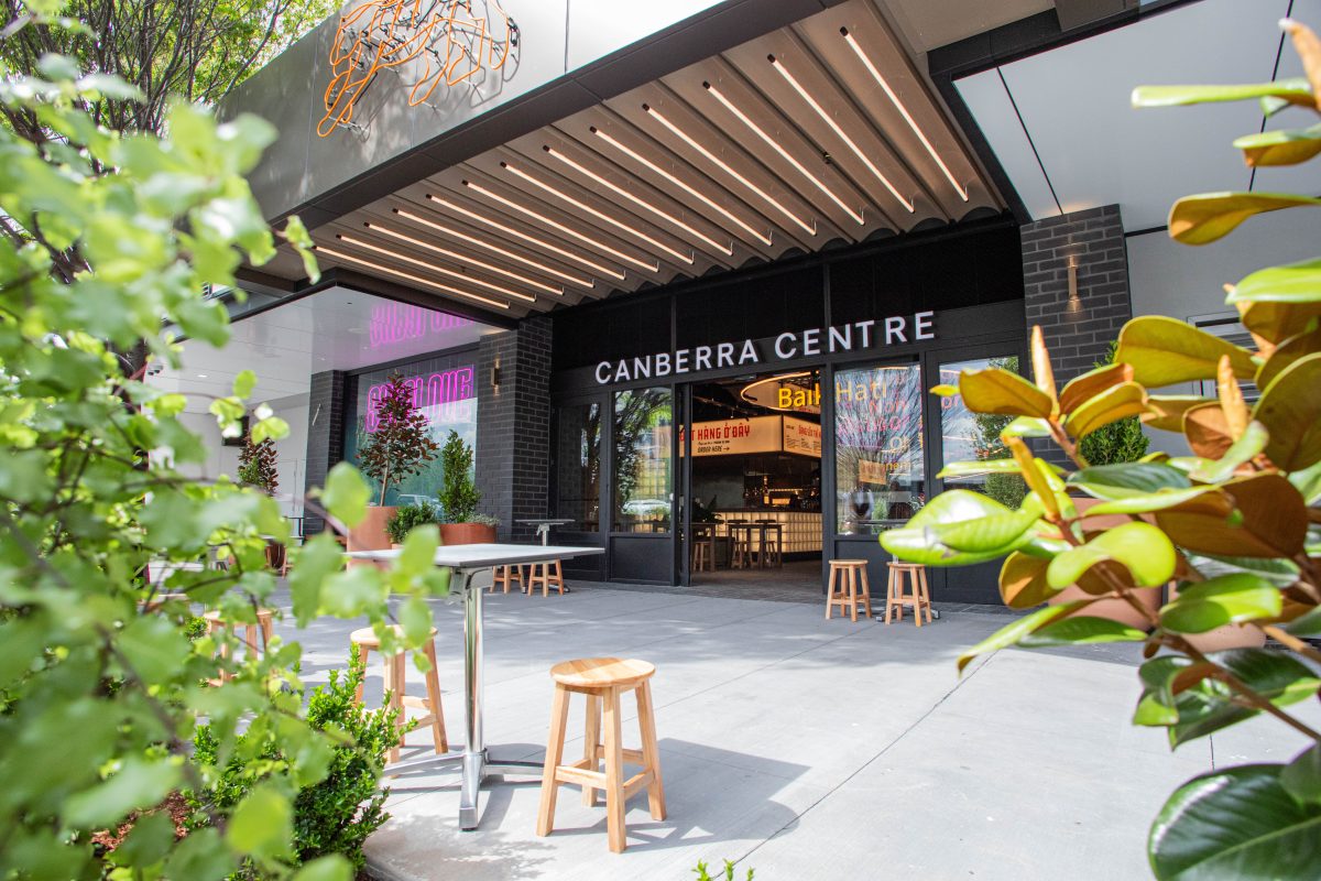 Exterior shot with Canberra centre sign, plants, tables and stools and a glimpse inside showing a food stall.