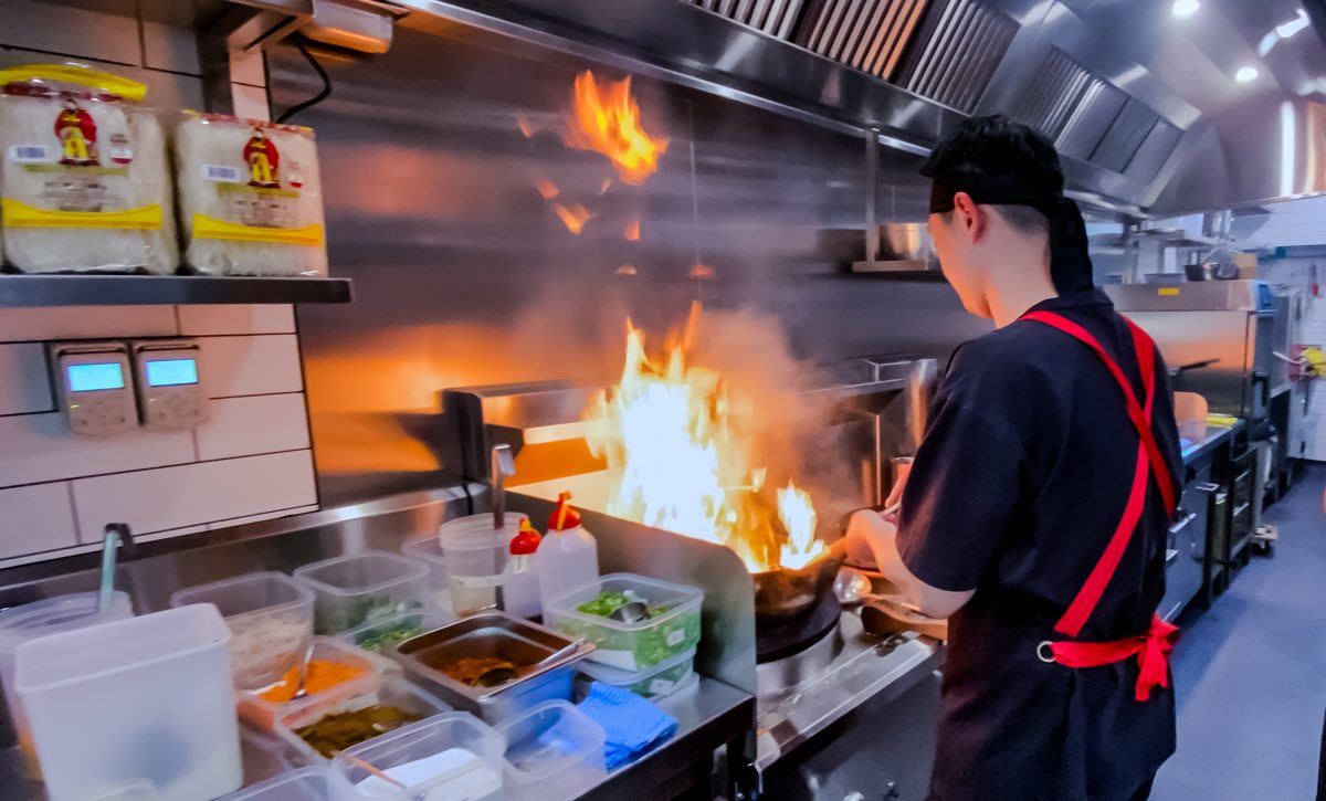 Chef in black with a fiery wok in a stainless steel kitchen.