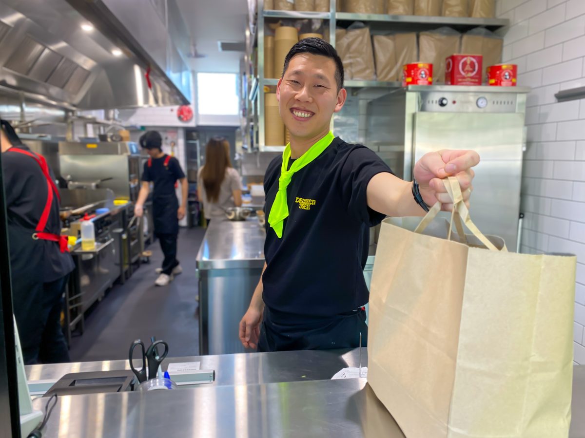 Jiwoo wears a black Drunken Tiger branded shirt and yellow necktie and smiles as he hands a paper bag across the counter.