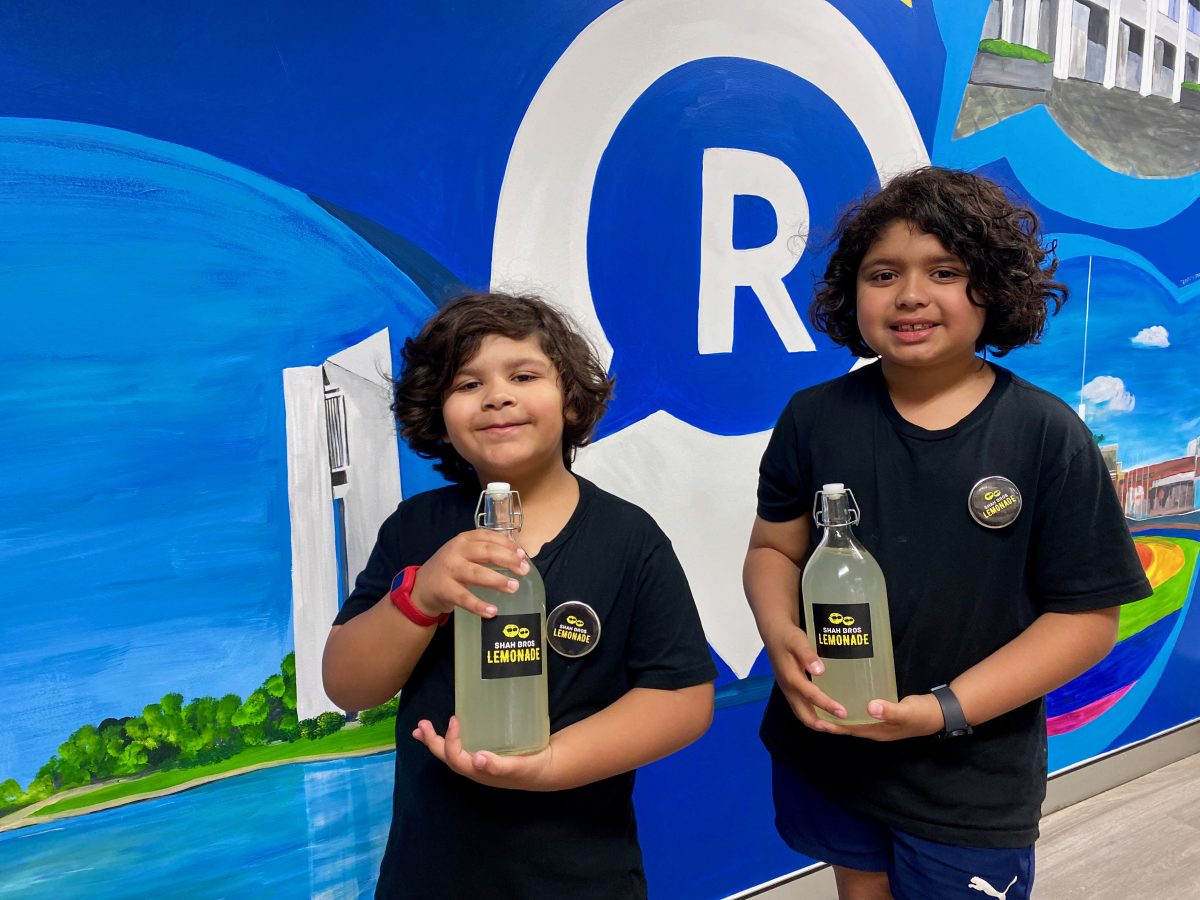 Two young boys stand in front of a mural with the Region Media logo. They smile and hold bottles of lemonade that say 'Shah Bros Lemonade'.