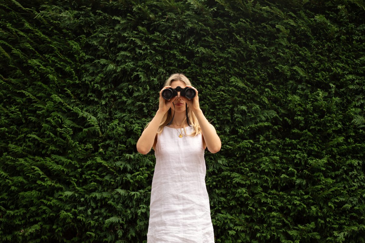 Long haired woman in white stands in front of hedge and holds binoculars.