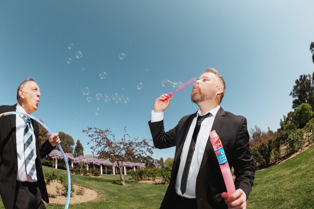 Two men in suits blow bubbles in a garden, photo it taken with a fish eye lens.