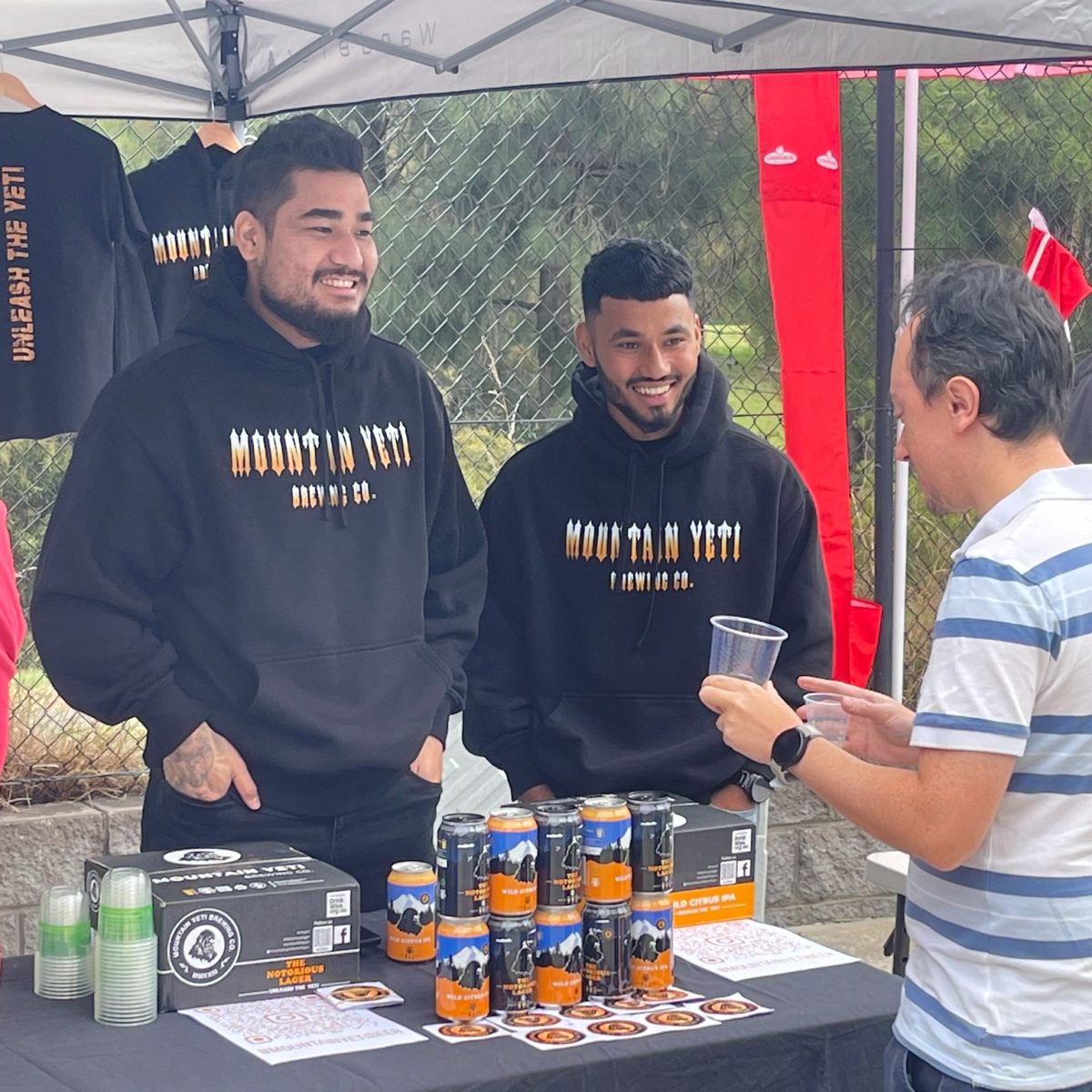 Two young Nepalese men wear Mountain Yeti Brewing Co branded black hoodies and serve beer to a customer at a market stall.