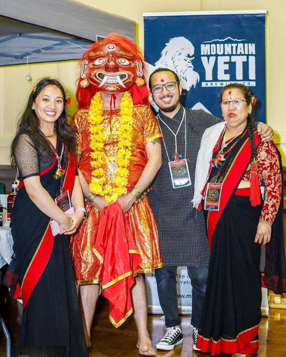 A figure wearing traditional 'lakhey' mask stands with three other people in Nepalese dress in front of Mountain Yeti Banner.