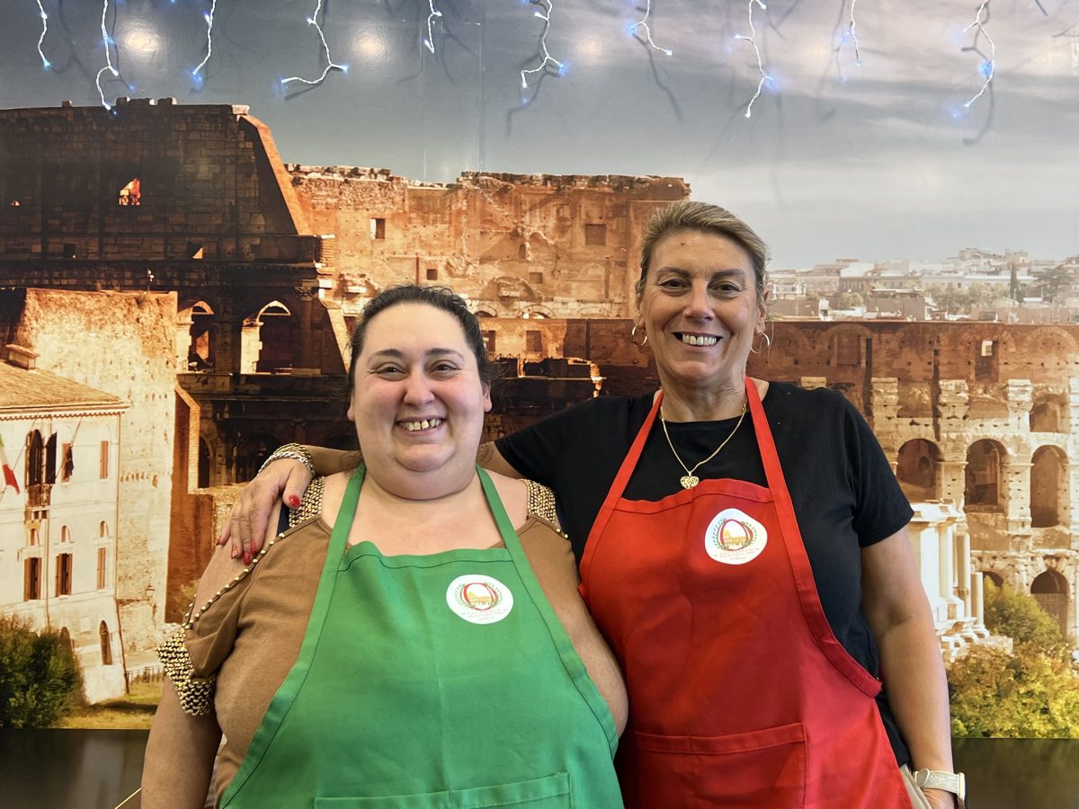 Sonya and Mariangela standing side by side with their aprons on in front of a colosseum wallpaper.
