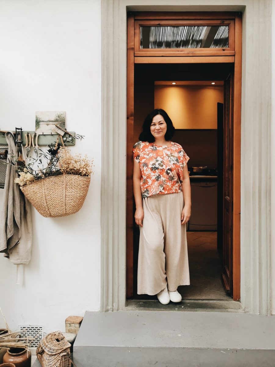Emiko stands in a doorway with a basket hanging next to her. Emiko has shoulder length dark hair and wears a loose colourful blouse and linen slacks.