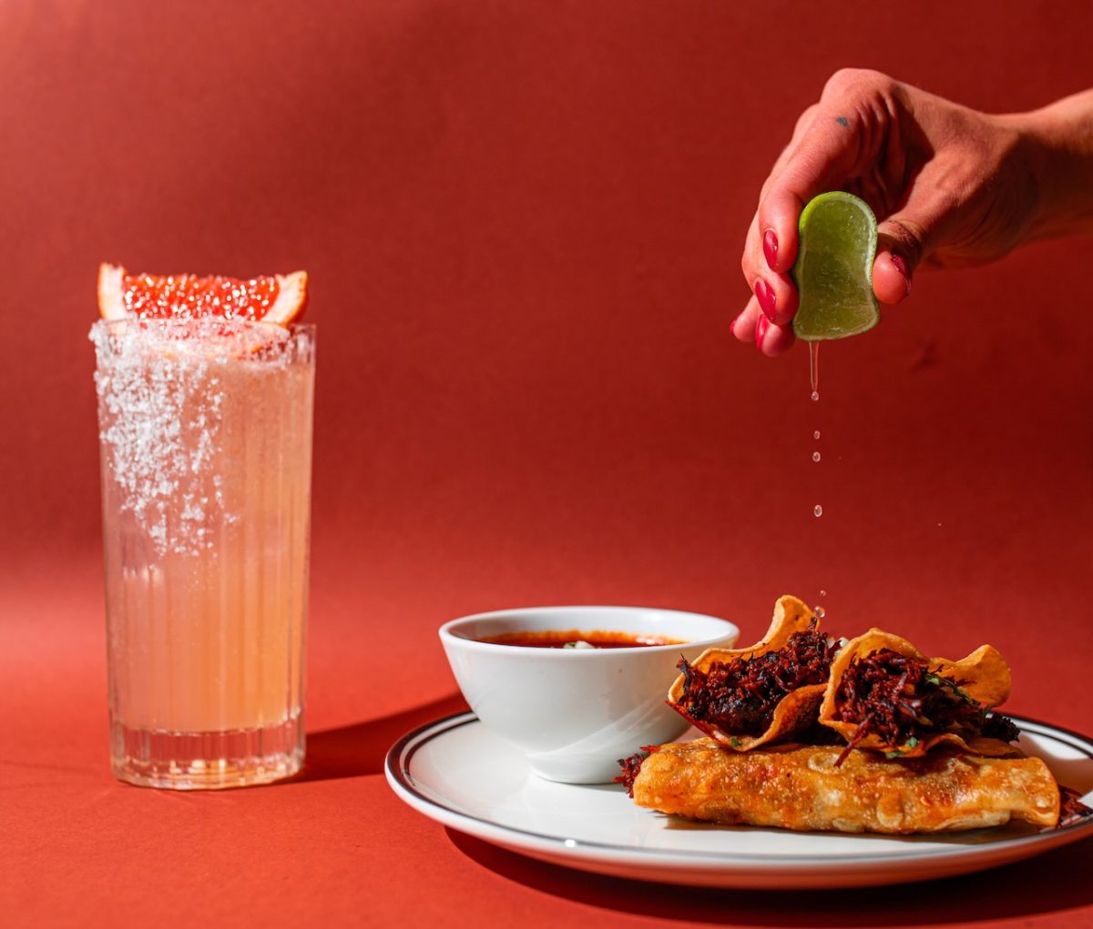 person squeezes lime onto crispy looking tacos. Red background and highball drink in background.