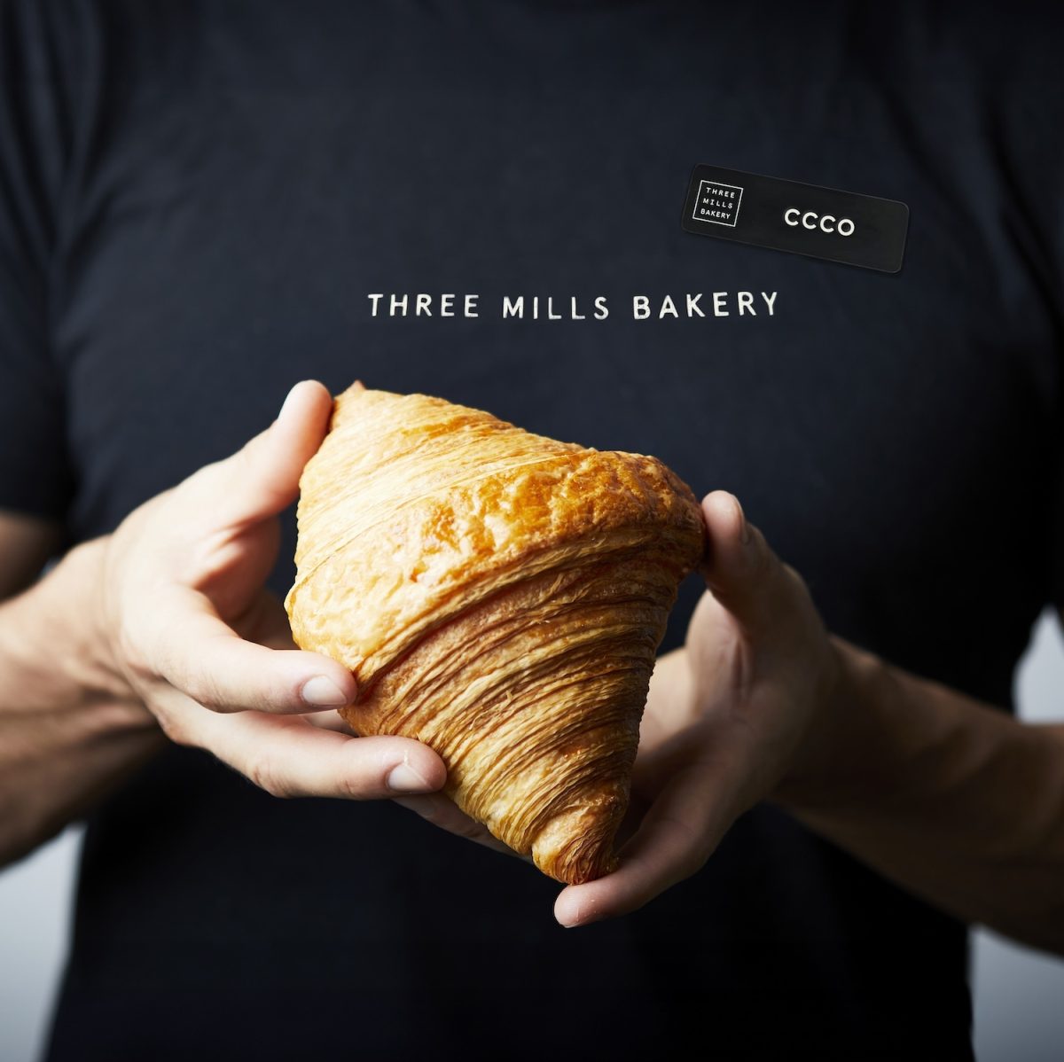 White hands hold a croissant in front of black shirt with white Three Mills Bakery text.