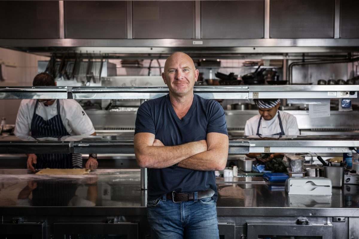 Matt Moran wears blue, crosses arms, in front of kitchen where chefs are working.