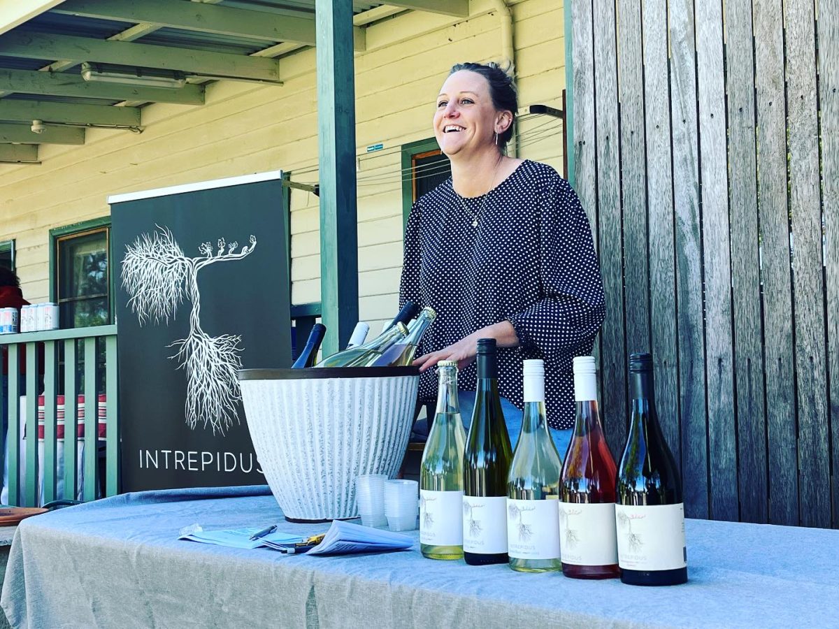 Chrissie stands behind a table with six bottles of wine, in the background a banner with the Intrepidus logo.