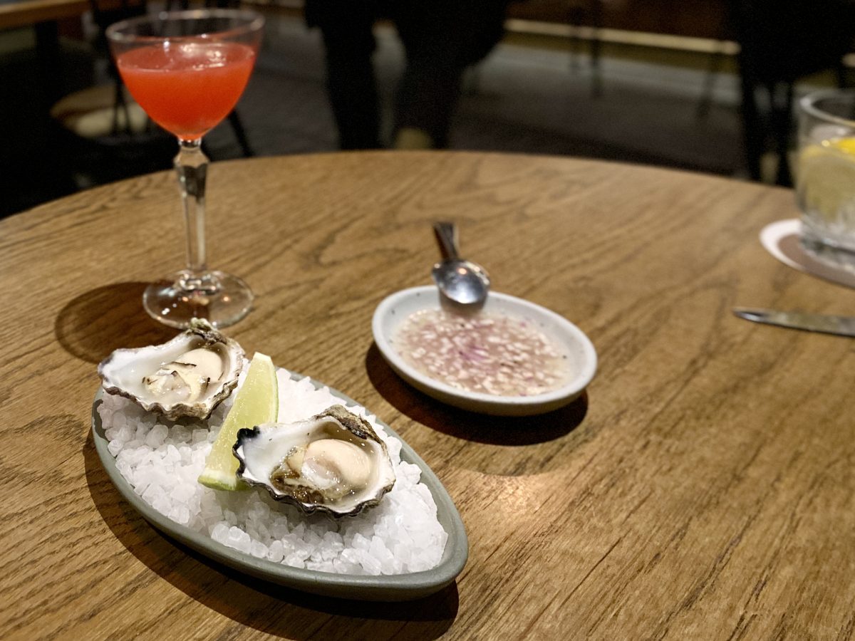 Two oysters on rocksalt with a ramekin of dressing and a pink cocktail in the background.