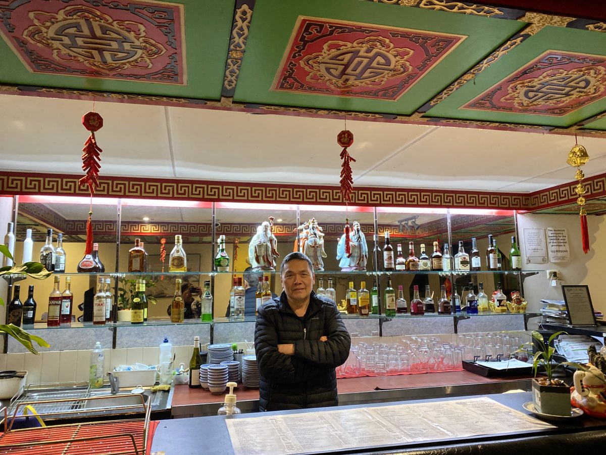 Older man stands with folded arms in from of bar with stylised Chinese decorations.