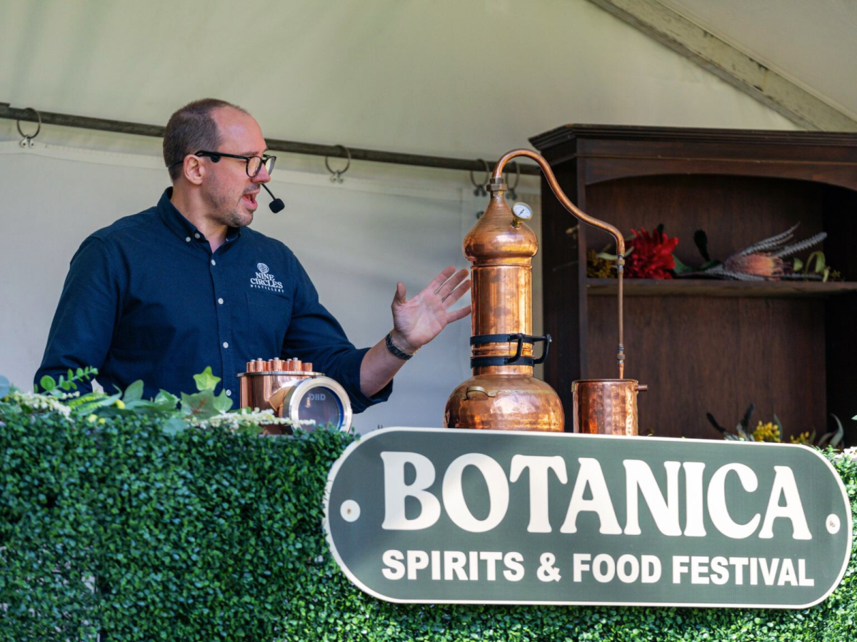 Man demonstrates workings of a copper still at an event signposted "Botanica Spirits and Food Festival". 