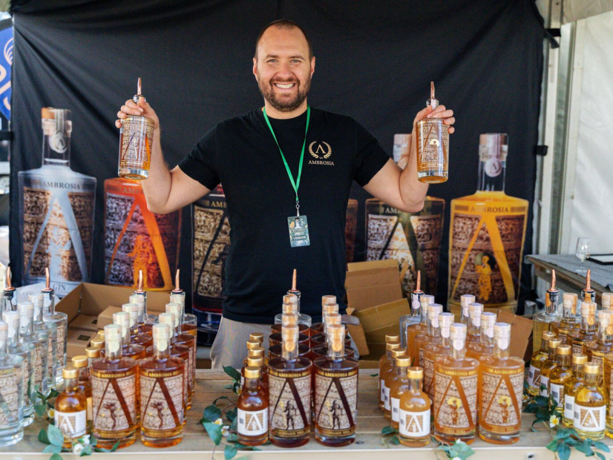 Man smiles holding two bottles of spirits with dozens of other bottles in the foreground