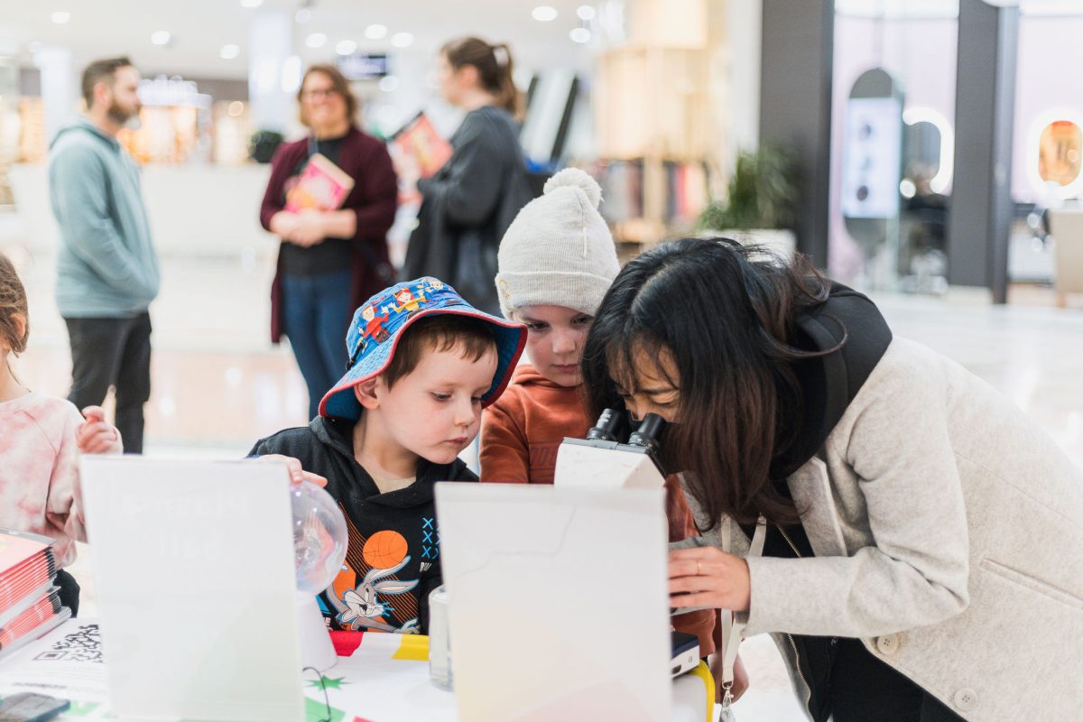 kids standing near a table looking at someone looking through a microscope