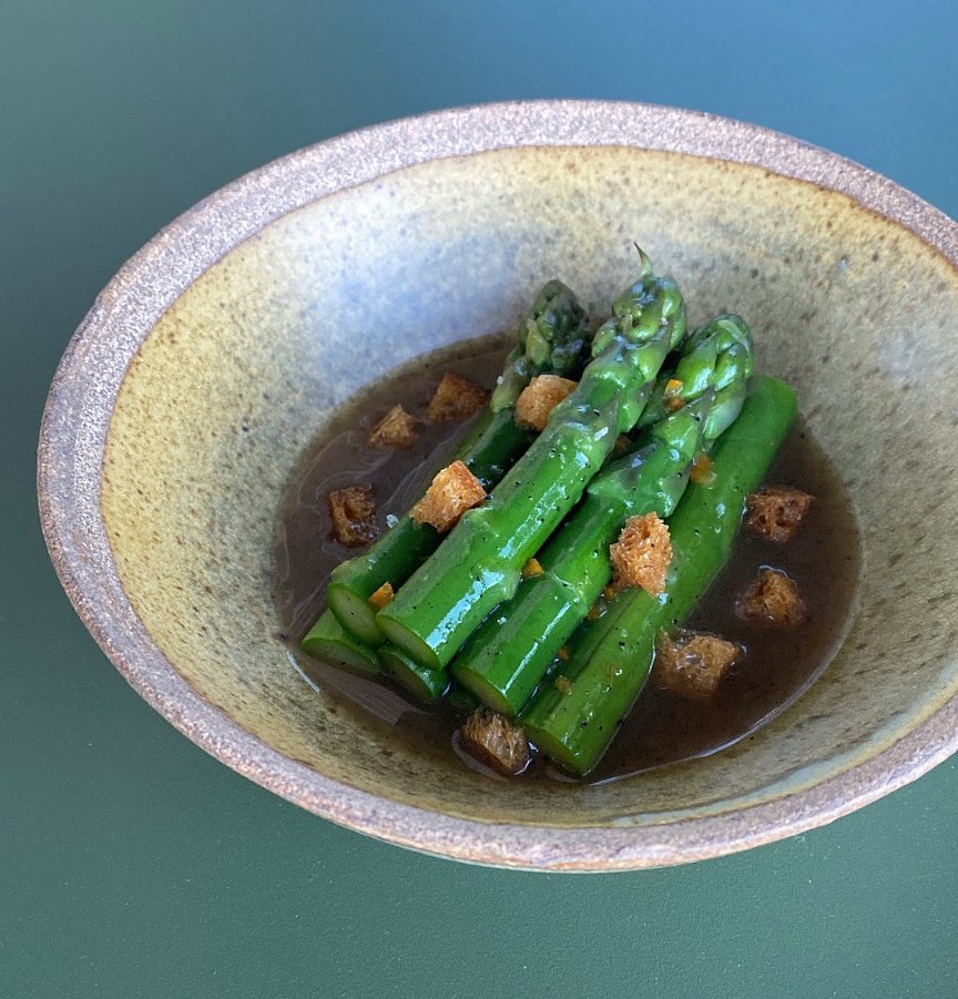 An earthenware bowl filled with green asparagus stems in a dark broth, scattered with croutons.