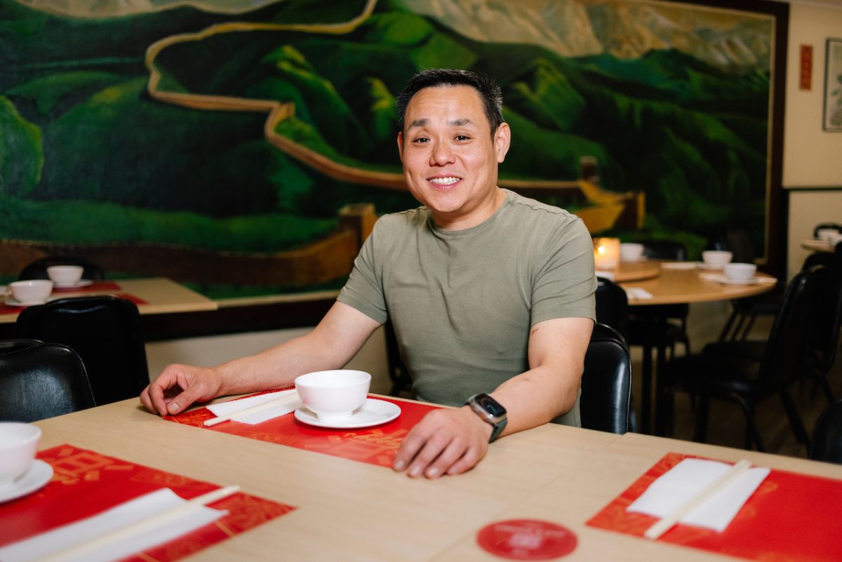 Gavin sits at a table laid with small bowls and chopsticks in front of a large mural depicting the great wall of China.