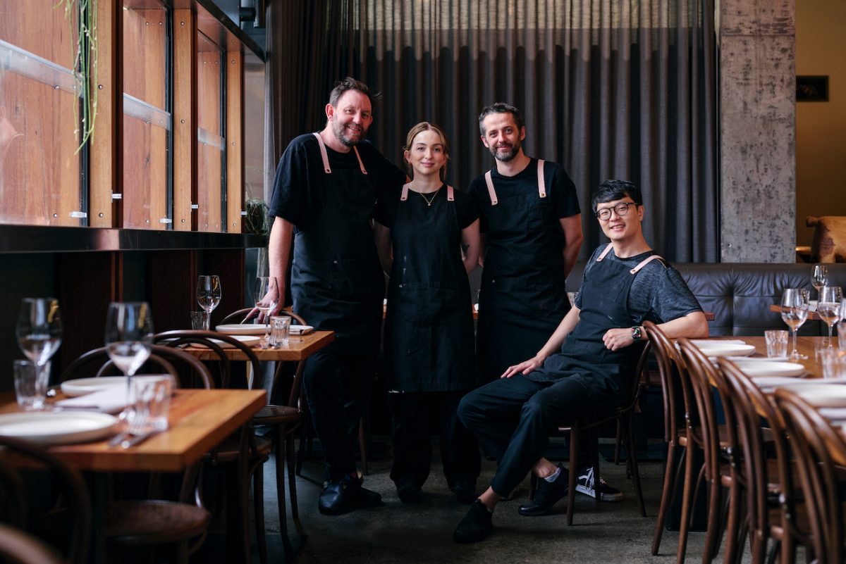 Four people in aprons pose in restaurant.