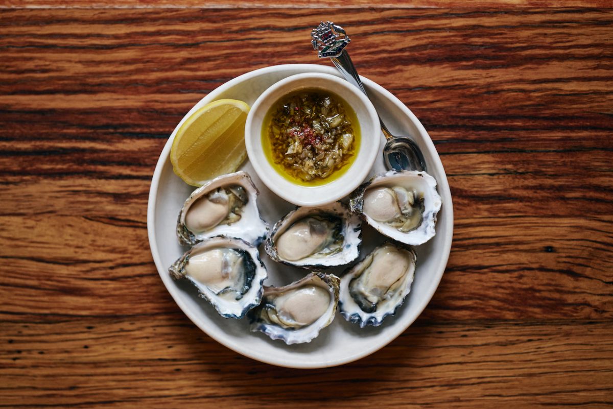 A plate of 6 plump oysters on a wooden table with a bowl of vinaigrette and cheek of lemon.