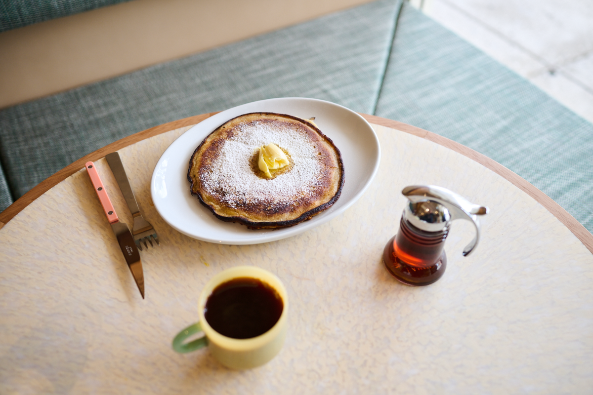 Oval plate with large pancake topped with butter and icing sugar sits on a table with a black coffee and jug of maple syrup.