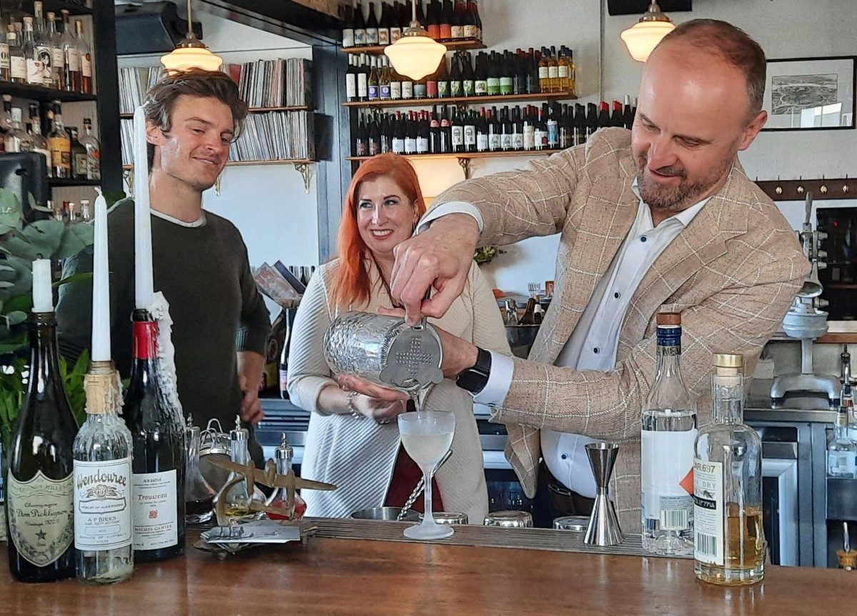 A man pours a cocktail in a bar while two others watch on