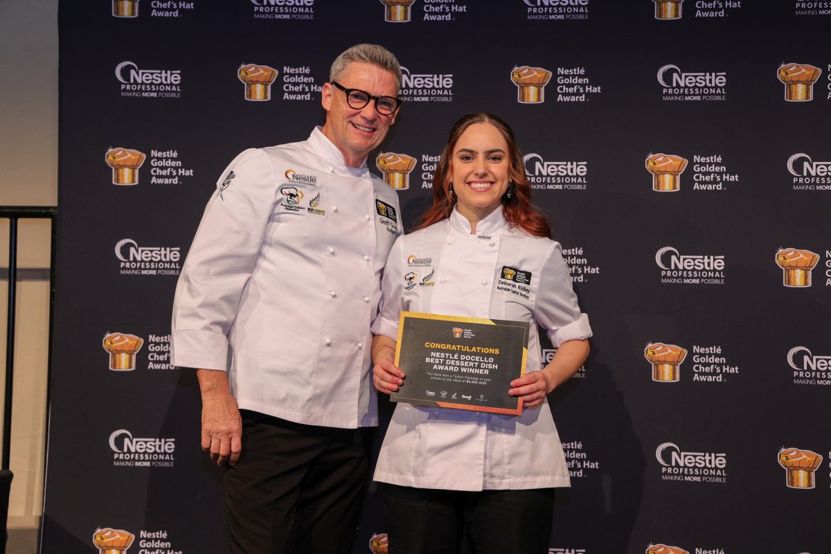 An older man in glasses and young woman holding a certificate both wear chef uniforms and smile in front of branded backdrop. 